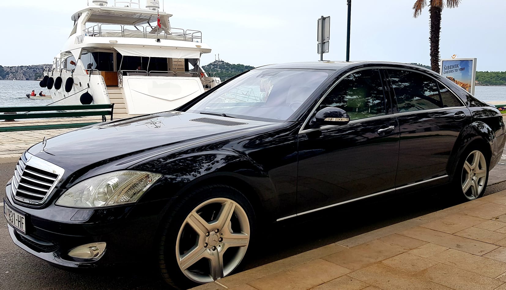 Black Mercedes S-Class luxury sedan parked beside a superyacht in Šibenik harbor — Cipollina VIP hotel and marina transfer service
